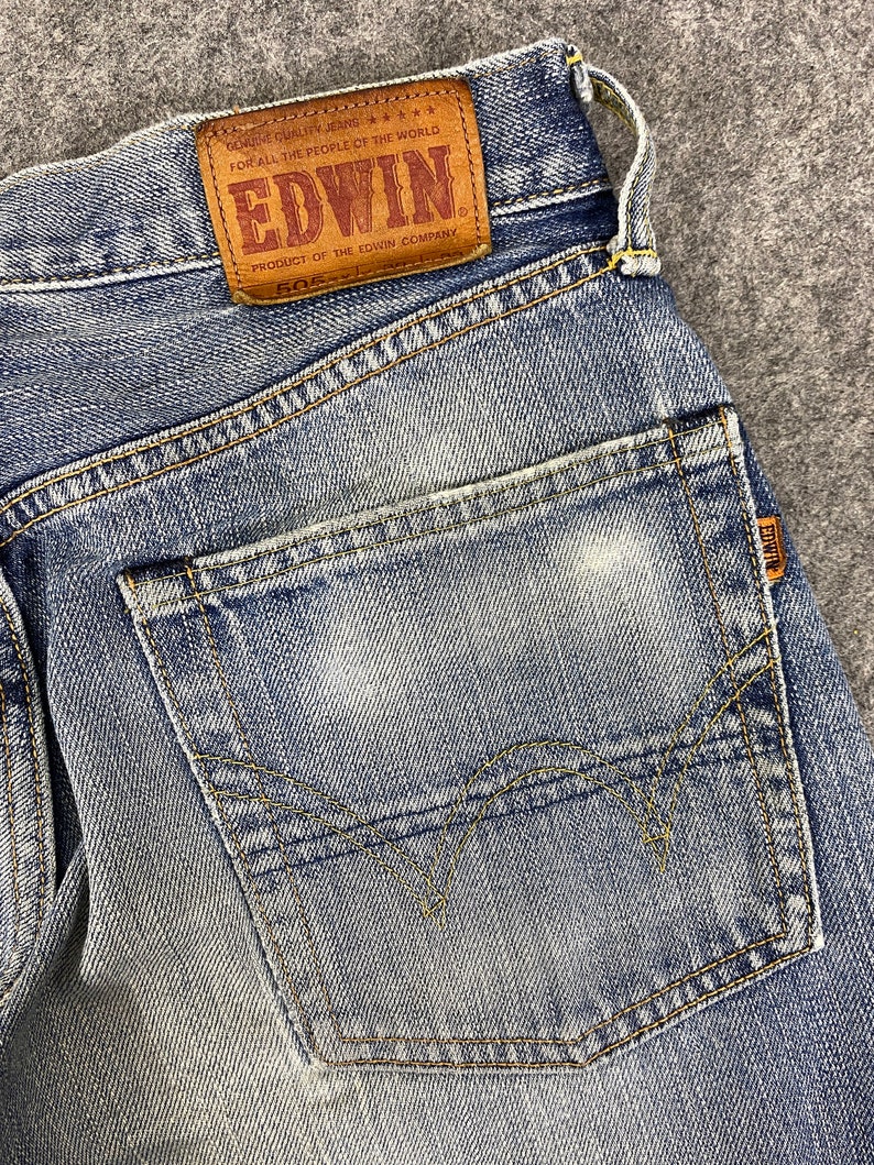 May include: Close-up of light blue denim jeans with a faded wash. The back pocket is visible, along with the Edwin brand leather patch. The patch reads "EDWIN" and "505".