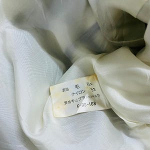 K&ouml;nnte beinhalten: Nahaufnahme des Inneren eines Kleidungsst&uuml;cks mit cremefarbenem Futter. Ein rechteckiges Etikett zeigt japanischen Text mit Prozentangaben f&uuml;r Woll- und Nylonanteil. Das Etikett enth&auml;lt auch den Text "C-OS-168".