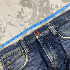 May include: Dark blue denim jeans featuring a distressed finish, yellow contrast stitching, and a red metal button at the waist. A blue measuring tape is positioned along the waistband, indicating a measurement of approximately 18 inches across.