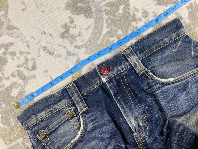 May include: Dark blue denim jeans featuring a distressed finish, yellow contrast stitching, and a red metal button at the waist. A blue measuring tape is positioned along the waistband, indicating a measurement of approximately 18 inches across.