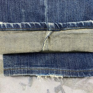 May include: Close-up of the hem of a pair of blue denim jeans. The jeans are folded up at the bottom, revealing the inside of the fabric. The denim is a dark blue color and has a faded look.