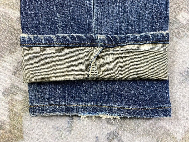 May include: Close-up of the hem of a pair of blue denim jeans. The jeans are folded up at the bottom, revealing the inside of the fabric. The denim is a dark blue color and has a faded look.