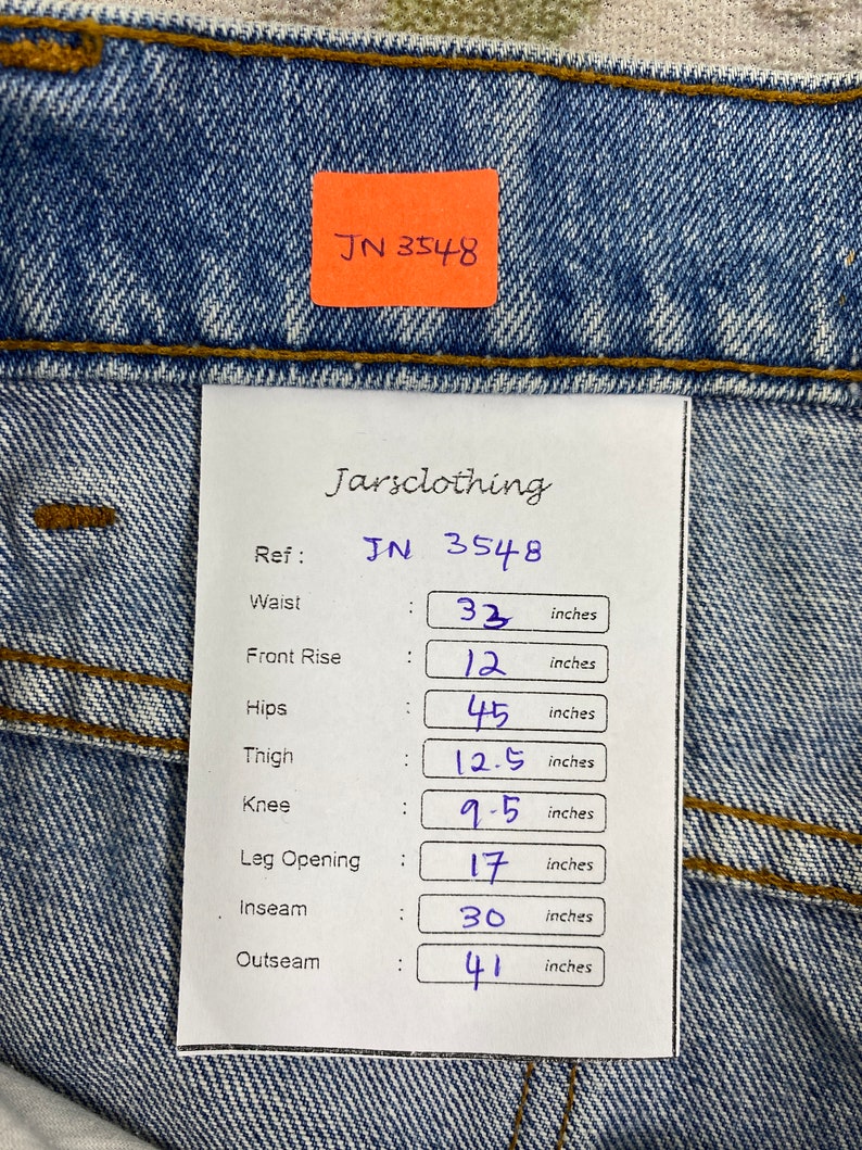 K&ouml;nnte beinhalten: Nahaufnahme einer blauen Jeans mit einem Etikett mit der Aufschrift "JN 3548". Die Taille misst 83,8 cm, die vordere Leibh&ouml;he 30,5 cm, die H&uuml;fte 114,3 cm, der Oberschenkel 31,8 cm, das Knie 24,1 cm, die Bein&ouml;ffnung 43,2 cm, die Innenbeinl&auml;nge 76,2 cm und die Au&szlig;enbeinl&auml;nge 104,1 cm.