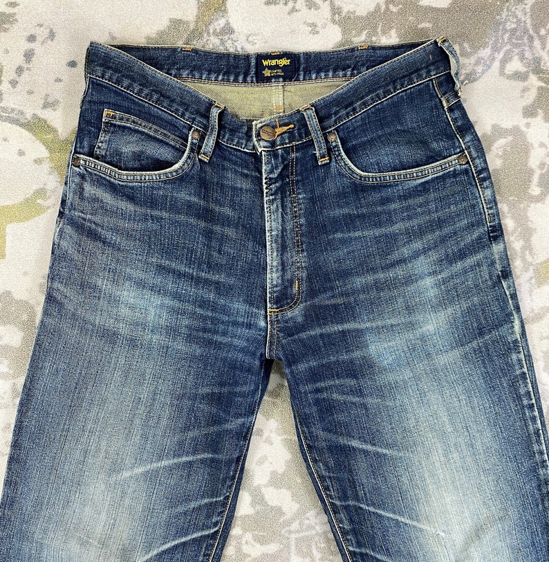 May include: A pair of blue denim jeans with a faded look. The jeans have a classic five-pocket style and a Wrangler brand label on the back waistband.
