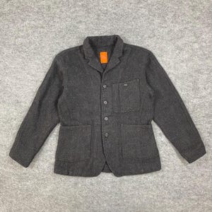 Puede incluir: Un blazer gris oscuro con un diseño clásico. La chaqueta presenta un cuello con solapas, tres botones, dos bolsillos delanteros y dos bolsillos en el pecho. El blazer tiene mangas largas y un corte entallado, adecuado para diversas ocasiones.