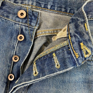 May include: Close-up of blue denim jeans with visible button closure and yellow stitching. The jeans have a faded wash and show signs of wear. The buttons are metallic with the brand name "EDWIN" visible.