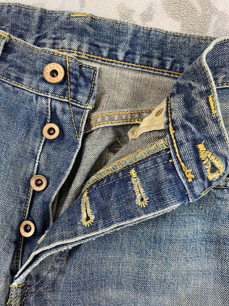May include: Close-up of blue denim jeans with visible button closure and yellow stitching. The jeans have a faded wash and show signs of wear. The buttons are metallic with the brand name "EDWIN" visible.