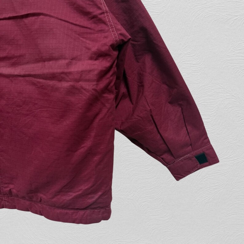 May include: A burgundy jacket with a textured, ribbed fabric. The jacket features a buttoned cuff and a small, dark green patch on the sleeve. The garment is shown against a white background.