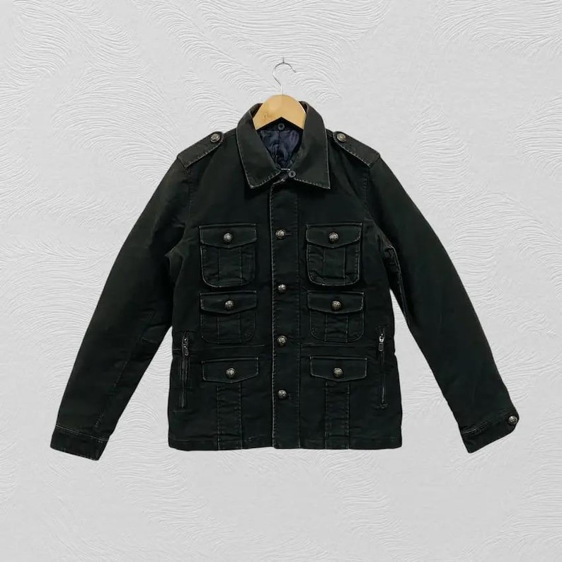 May include: Black jacket with multiple front pockets and button closures. The jacket has a collared neckline and long sleeves. It is hanging on a wooden hanger against a white background.
