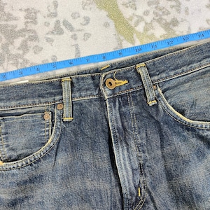 May include: Close-up of blue denim jeans with a button closure and front pockets. A blue measuring tape shows the waist size. The jeans have a worn, distressed look.