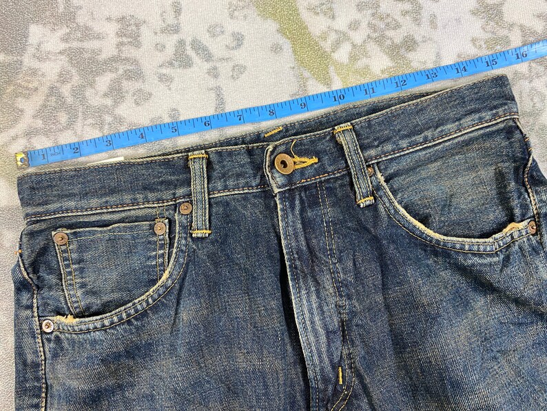 May include: Close-up of blue denim jeans with a button closure and front pockets. A blue measuring tape shows the waist size. The jeans have a worn, distressed look.