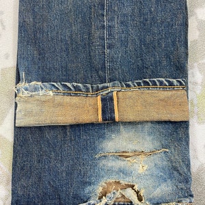 May include: Close-up of distressed blue denim jeans. The jeans have a faded wash, a torn section near the knee, and a folded cuff revealing a lighter, tan-colored interior. The stitching is visible, and the fabric shows signs of wear.