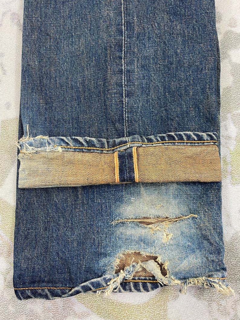 May include: Close-up of distressed blue denim jeans. The jeans have a faded wash, a torn section near the knee, and a folded cuff revealing a lighter, tan-colored interior. The stitching is visible, and the fabric shows signs of wear.