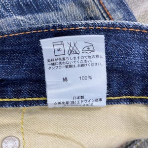 May include: A pair of blue denim jeans featuring a white interior care label with Japanese text, laundry symbols, and a 100% cotton composition. The jeans have visible orange stitching along the waistband and a metallic button with embossed text.