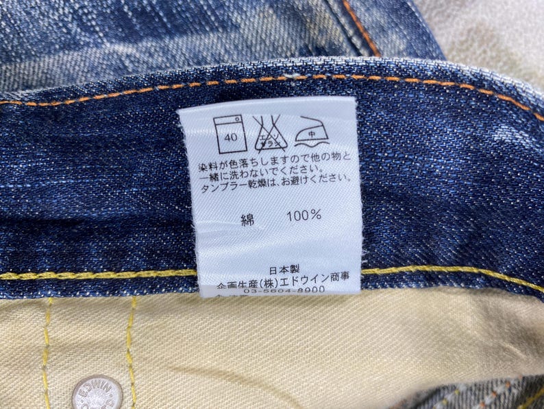 May include: A pair of blue denim jeans featuring a white interior care label with Japanese text, laundry symbols, and a 100% cotton composition. The jeans have visible orange stitching along the waistband and a metallic button with embossed text.