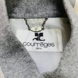 K&ouml;nnte beinhalten: Nahaufnahme eines grauen Wollmantelkragens mit wei&szlig;em Futter. Der Mantel hat ein Etikett mit dem Markennamen "Courr&egrave;ges Paris" in schwarzer Schrift. Eine dekorative Kette mit einem wolkenf&ouml;rmigen Anh&auml;nger ist am Kragen befestigt.