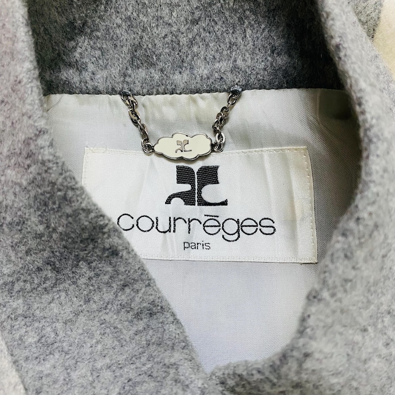 K&ouml;nnte beinhalten: Nahaufnahme eines grauen Wollmantelkragens mit wei&szlig;em Futter. Der Mantel hat ein Etikett mit dem Markennamen "Courr&egrave;ges Paris" in schwarzer Schrift. Eine dekorative Kette mit einem wolkenf&ouml;rmigen Anh&auml;nger ist am Kragen befestigt.