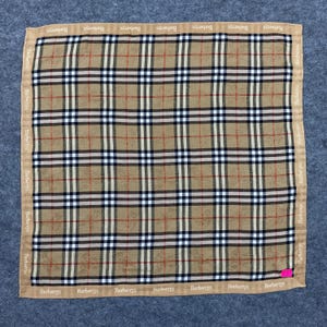 May include: A square scarf with a classic plaid pattern in tan, black, white, and red. The border features the brand name "Burberrys" in a repeating pattern. The scarf is made of a soft fabric.