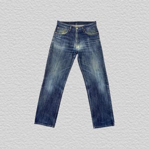 Könnte beinhalten: Eine blaue Jeans aus Denim mit klassischem Fünf-Taschen-Design, Knopfleiste und einem verwaschenen Effekt an den Oberschenkeln und am Gesäß. Die Hose hat einen geradlinigen Schnitt mit sichtbaren Nähten und metallischen Nieten.
