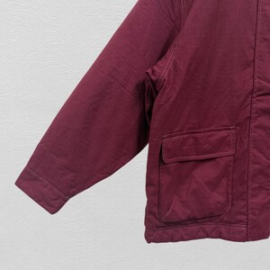 May include: A maroon jacket with a textured fabric and two square pockets. The jacket has a long sleeve and a visible seam running down the side. The background is a light gray.