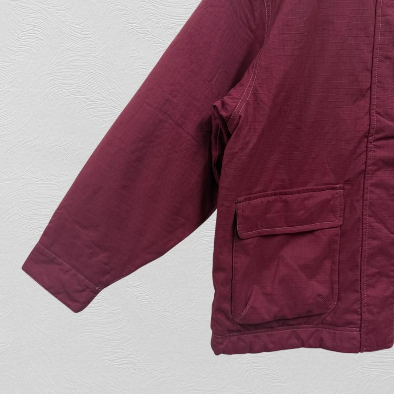 May include: A maroon jacket with a textured fabric and two square pockets. The jacket has a long sleeve and a visible seam running down the side. The background is a light gray.