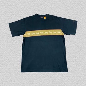 Pode incluir: Camiseta azul marinho escuro com uma faixa horizontal bege no peito. A faixa apresenta um padr&atilde;o repetitivo de silhuetas de elefantes brancos. A camiseta tem mangas curtas e gola redonda.