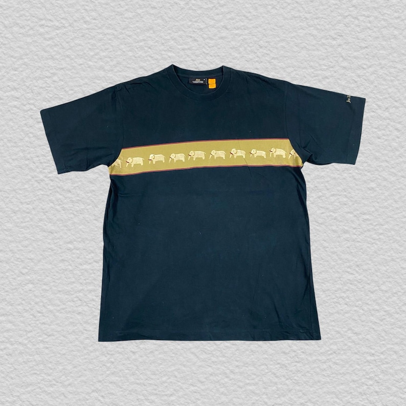 Pode incluir: Camiseta azul marinho escuro com uma faixa horizontal bege no peito. A faixa apresenta um padr&atilde;o repetitivo de silhuetas de elefantes brancos. A camiseta tem mangas curtas e gola redonda.