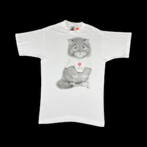 May include: White t-shirt featuring a detailed illustration of a gray cat wearing a shirt that says "I CATS" with a pink heart. The cat is the central focus, rendered in a realistic style. The shirt is laid flat on a black surface.