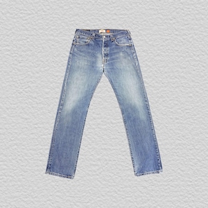 May include: A pair of medium-wash blue denim jeans featuring a classic five-pocket design, a button fly closure, copper-toned rivets, and a tan leather-like brand patch located on the back waistband. The denim has a faded finish throughout.