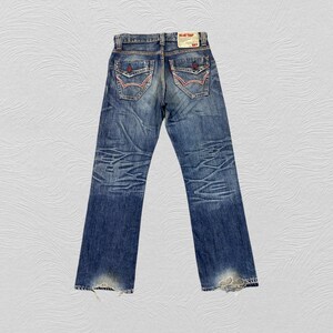 May include: Blue denim jeans featuring a faded wash, whiskering patterns on the thighs, and two back pockets with red and white stitching details. A rectangular white label at the waistband displays the text BLUE TRIP and 503 in red ink.