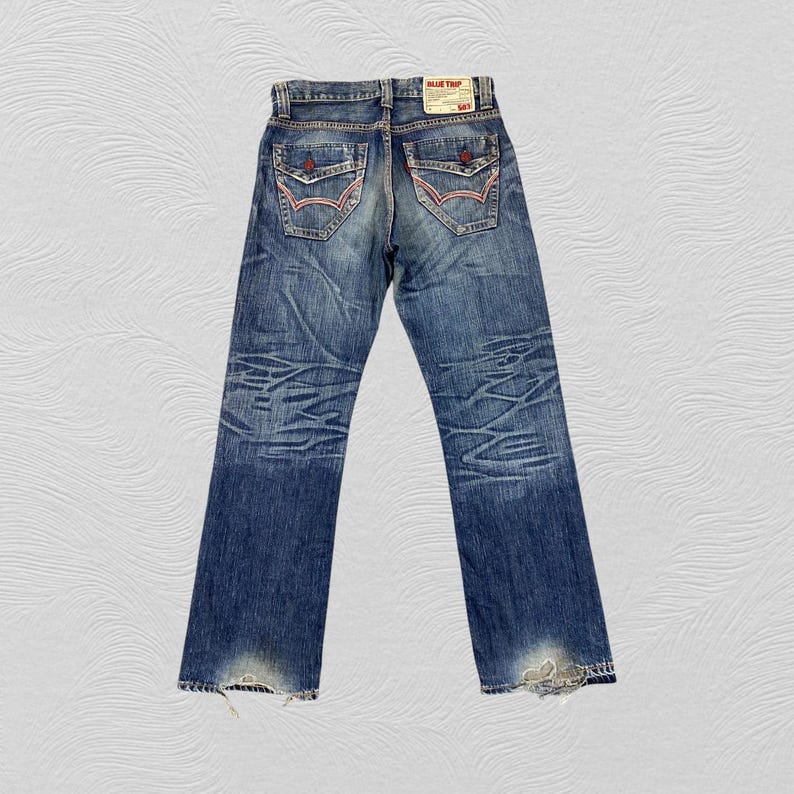 May include: Blue denim jeans featuring a faded wash, whiskering patterns on the thighs, and two back pockets with red and white stitching details. A rectangular white label at the waistband displays the text BLUE TRIP and 503 in red ink.