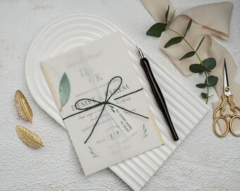 May include: A wedding invitation with a watercolor floral design and a black ribbon tied around it. The invitation is on a white surface with a gold pen, a pair of gold scissors, and a sprig of eucalyptus.