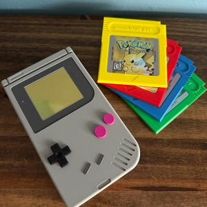 Puede incluir: Una consola de videojuegos portátil de color gris claro con un mando direccional negro, dos botones rosas y dos botones pequeños grises. Junto a ella hay cuatro cartuchos de juego coloridos en amarillo, rojo, azul y verde, con el amarillo etiquetado Pokemon.