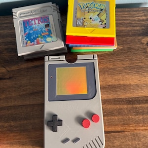 May include: A gray Nintendo Game Boy console and game cartridges. The cartridges include Tetris and Pokémon Special Pikachu Edition. The console has a directional pad, red buttons, and a screen. The cartridges are stacked on top of each other.