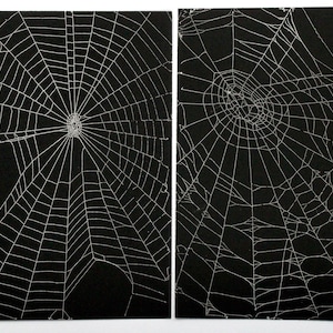Op de afbeelding: Twee rechthoekige prints met witte spinnenwebpatronen tegen een effen zwarte achtergrond. Elke print toont een gedetailleerde, ingewikkelde radiale webstructuur met fijne lijnen die vanuit een centraal punt uitwaaieren voor een contrastrijk ontwerp.