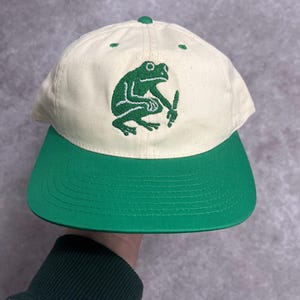 Könnte beinhalten: Cremefarbene Baseballkappe mit grünem Schirm und grünem Frosch-Motiv. Der Frosch ist auf der Vorderseite der Kappe gestickt und hält ein kleines Objekt. Die Kappe hat einen gebogenen Schirm und einen Knopf oben.