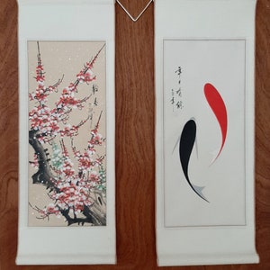 May include: Two framed scrolls featuring traditional Asian artwork. The left scroll depicts a delicate painting of a blossoming tree branch with pink flowers. The right scroll shows two stylized fish, one red and one black, swimming in a circle. The scrolls are hung on a wooden wall with a decorative metal hanger.