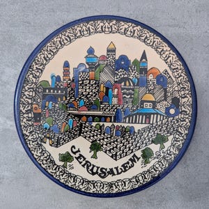 May include: A round ceramic decorative plate featuring a hand-painted illustration of the city of Jerusalem. The design includes colorful buildings, domes, and stone walls with the word JERUSALEM printed at the bottom, framed by a dark blue border.