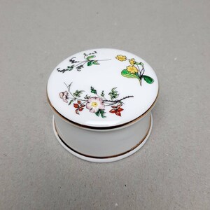 Puede incluir: Una caja de porcelana blanca con tapa redonda. La tapa está decorada con un diseño floral con flores rosas, amarillas y verdes. La caja tiene un borde dorado.