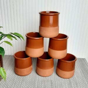 May include: Six terracotta ceramic pots stacked in a pyramid. Each pot is cylindrical with a rounded rim and a two-tone design. The top half is a glossy brown, and the bottom half is a matte peach colour. The pots are for decorative or practical use.