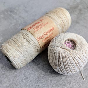 May include: Two spools of natural-coloured thread. One is cylindrical with a paper label featuring red text, and the other is a ball of thread with a circular label. The thread appears to be made of a natural fibre.
