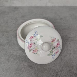 Puede incluir: Una caja de cerámica blanca para baratijas con una tapa con un diseño floral en rosa, azul y verde. La caja tiene un borde rosa y un pequeño pomo redondo en la tapa. La caja abierta está sobre una superficie gris.