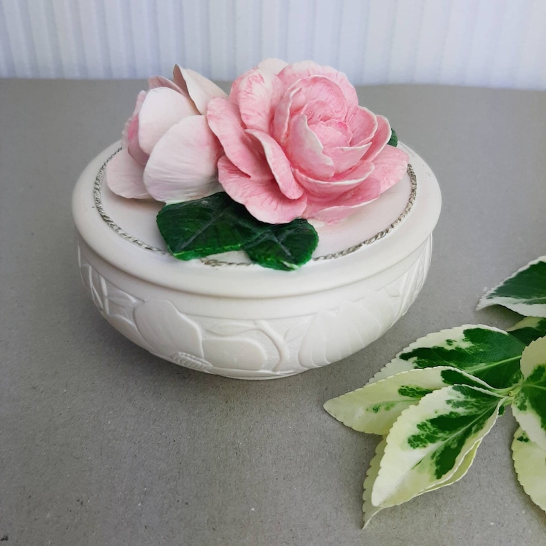 Puede incluir: Caja de cer&aacute;mica blanca con un dise&ntilde;o floral. La tapa presenta dos rosas rosadas con hojas verdes.
