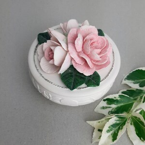 Puede incluir: Una caja de cer&aacute;mica blanca con un dise&ntilde;o floral. La tapa presenta tres rosas rosadas con hojas verdes.