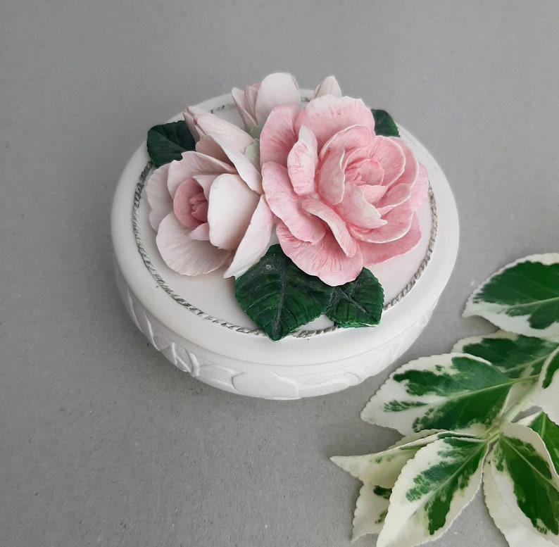 Puede incluir: Una caja de cer&aacute;mica blanca con un dise&ntilde;o floral. La tapa presenta tres rosas rosadas con hojas verdes.