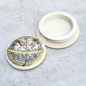 May include: A white ceramic trinket box with a lid featuring a vintage celestial map with constellations and a compass. The lid is decorated with a yellow rim and the words "HORIZON" in black.