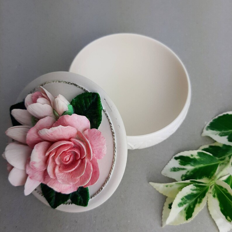 Puede incluir: Caja de cer&aacute;mica blanca con tapa con una rosa rosa y una flor blanca con hojas verdes. La caja est&aacute; abierta y el interior es visible.