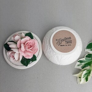 Puede incluir: Caja de cer&aacute;mica blanca con un dise&ntilde;o floral. La tapa presenta un grupo de rosas rosas y hojas verdes. La parte inferior de la caja tiene el texto "Handcast Designs Limited Made in U.K."