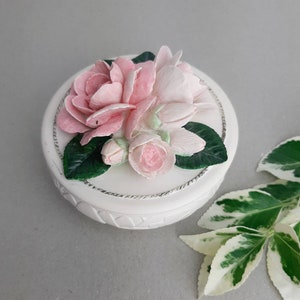 Puede incluir: Caja de cer&aacute;mica blanca con un dise&ntilde;o floral. La tapa presenta un grupo de flores rosas y blancas con hojas verdes.