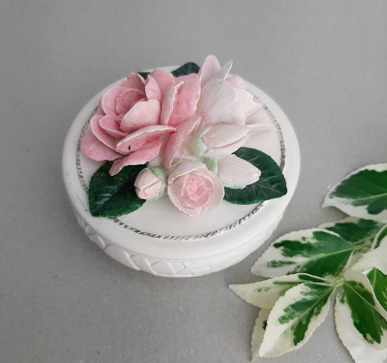 Puede incluir: Caja de cer&aacute;mica blanca con un dise&ntilde;o floral. La tapa presenta un grupo de flores rosas y blancas con hojas verdes.